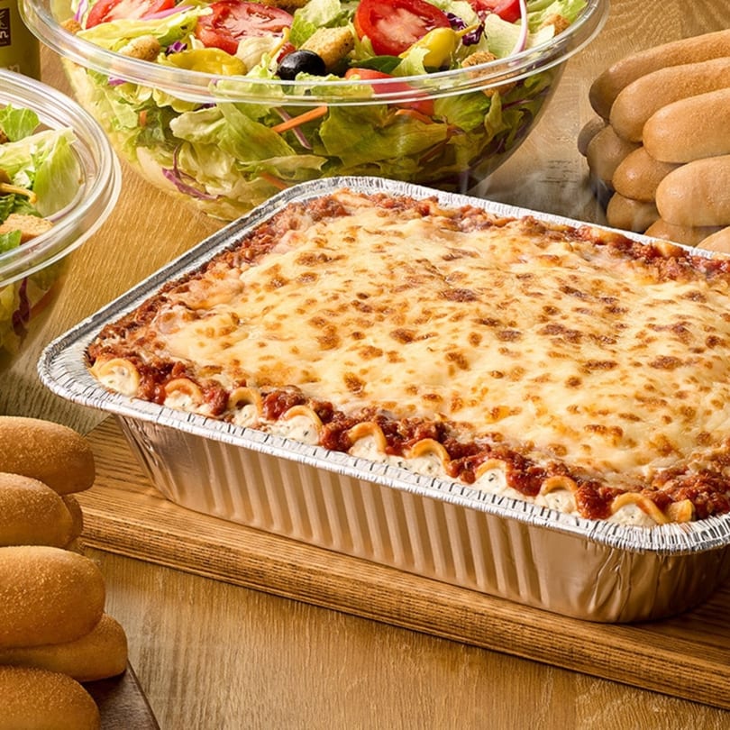 Baked lasagna in a foil pan, with two large bowls of salad and bread rolls on a wooden table.